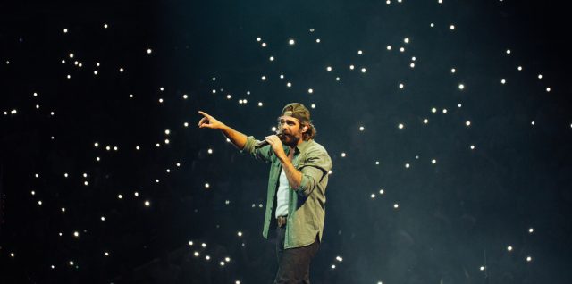 Thomas Rhett sings on a concert stage surrounded by fans holding phone lights