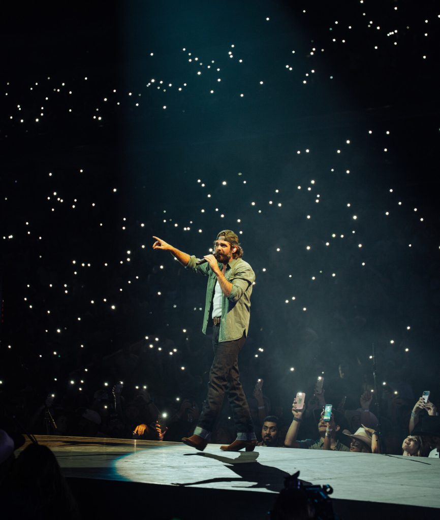 Thomas Rhett sings on a concert stage surrounded by fans holding phone lights